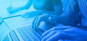 Two people are at a desk writing. A female is writing with a pen on notepad and a male is typing on a laptop.