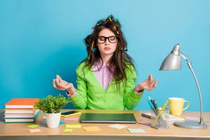 A woman with multiple pencils in her hair, sitting behind a desk, is trying to meditate.